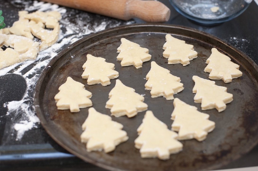 Ogräddade julgransformade pepparkakor på en bakplåt, med kavel och mjöl i bakgrunden.